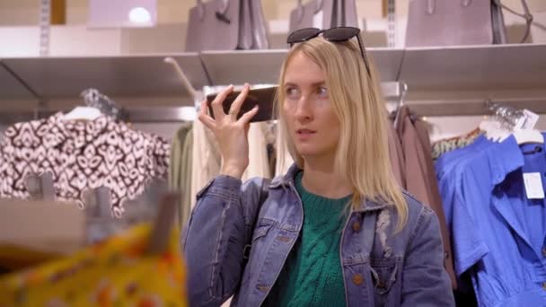 아름답고 Bulndinka 의류가게 센터에서 스마트폰을 그녀는 선택하고 메신저로 메시지를 듣는다 — 비디오