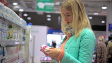 Young blonde beauty holds cotton pads in cosmetics department, and studies composition and manufacturer. Treasurer at grocery store inspects goods on shelves. Trading row of market.