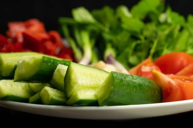 Siyah arka planda beyaz bir tabakta doğranmış taze otlar ve sebzeler. Salatalık, domates, dolmalık biber, yeşil soğan, horoz. Sebzelerden yararlı vitaminler
