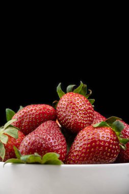 Siyah arka planda yeşil kuyruklu olgun kırmızı çilekleri beyaz bir tabakta yakın. Masada taze böğürtlenli tatlı. Dikey fotoğraf