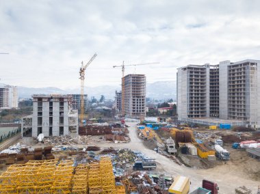 İnşaat alanının insansız hava aracı görüntüsü, çok katlı binalar, dağların arka planına karşı inşaat halinde. İnşaat makineleri, kazıcılar, çekici vinç