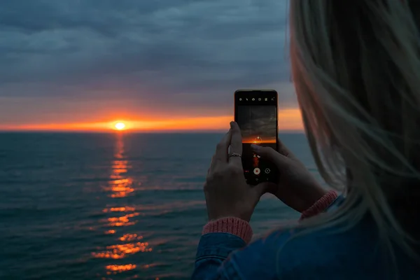 Bonde woman takes a video of the sun going over the horizon of the sea on a mobile phone, copy space. Shooting a sunset video on the sea using a mobile phone