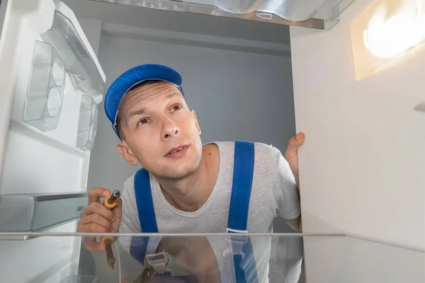 Male technician in overalls repairs a broken refrigerator with a screwdriver. Refrigerator repair concept. Photo from inside the refrigerator