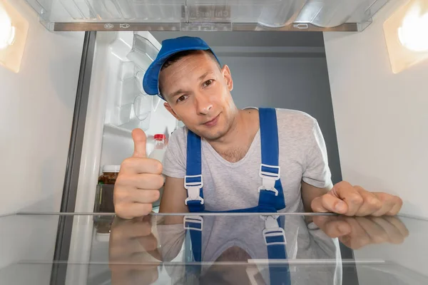 Portrait of a male repairman in uniform inside a refrigerator, showing a thumbs up sign. Concept of repair of household appliances. Photo from inside the refrigerator