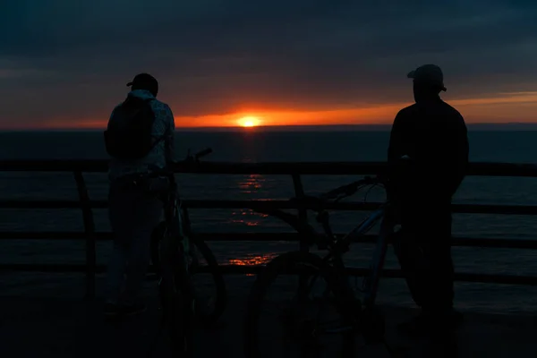 Siluetas Dos Hombres Con Bicicletas Muelle Observando Puesta Sol Mar — Foto de Stock