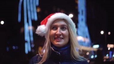 Makyajlı bir kadın portresi, kameraya gülümseyen ve gece veya akşam şehir ışıklarının arka planında Noel Baba şapkası takan. Gülümser, yeni yılı ya da Noel 'i kutlar..