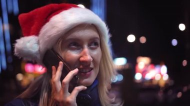 a close-up photo of a white woman chatting on her smartphone. A pretty woman in a Santa hat. A cute girl is talking on the phone and smiling against the background of city lights on Christmas Day.