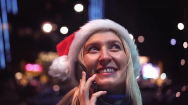 Beyaz bir kadının akıllı telefonuyla sohbet ederken çekilmiş yakın plan fotoğrafı. Noel Baba şapkalı güzel bir kadın. Güzel bir kız telefonda konuşuyor ve Noel 'de şehrin ışıklarına gülümsüyor..