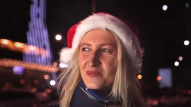 Portrait of woman with makeup, she yawns covering her mouth with her palm and smiles at camera in Santa hat against background of city lights at night. She is happy celebrating New Year or Christmas.