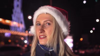 Makyajlı bir kadın portresi, kameraya gülümseyen ve gece veya akşam şehir ışıklarının arka planında Noel Baba şapkası takan. Gülümser, yeni yılı ya da Noel 'i kutlar..