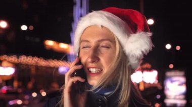 Beyaz bir kadının akıllı telefonuyla sohbet ederken çekilmiş yakın plan fotoğrafı. Noel Baba şapkalı güzel bir kadın. Güzel bir kız telefonda konuşuyor ve Noel 'de şehrin ışıklarına gülümsüyor..