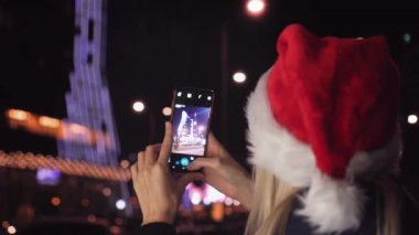 Noel Baba şapkalı gülümseyen bir kadın Noel arifesinde akıllı telefonuyla gece şehrinin fotoğrafını çekiyor. Mutlu bir kadın Noel arifesinde cep telefonuyla fotoğraf çeker..