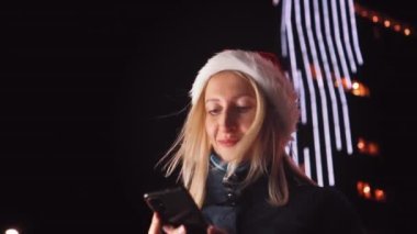 Noel Baba şapkalı güzel bir sarışının portresi, cep telefonuna mesaj yazıyor ve iyi haber alınca duygusal tepki veriyor. Şehir ışıklarının arka planındaki akıllı telefonlara bakar..