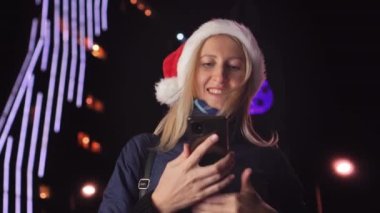 Noel Baba şapkalı güzel bir sarışının portresi, cep telefonuna mesaj yazıyor ve iyi haber alınca duygusal tepki veriyor. Şehir ışıklarının arka planındaki akıllı telefonlara bakar..