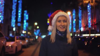 A beautiful woman in a Santa Claus hat is walking on a winter night. The street is decorated with garlands of flowers along the alley, decorated with lights on New Year's Eve or Christmas.