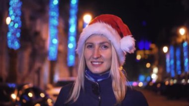 Makyajlı bir kadın portresi, kameraya gülümseyen ve gece veya akşam şehir ışıklarının arka planında Noel Baba şapkası takan. Gülümser, yeni yılı ya da Noel 'i kutlar..