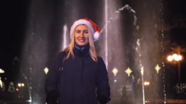 girl looks at the camera and smiles against background of the fountain on Christmas night or New Year's Eve. She's wearing a Santa Claus hat. night before Christmas. City lights in the background.