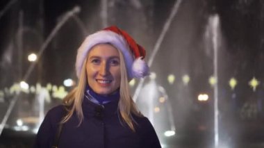 girl looks at the camera and smiles against background of the fountain on Christmas night or New Year's Eve. She's wearing a Santa Claus hat. night before Christmas. City lights in the background.