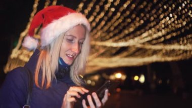 Noel Baba şapkalı güzel bir sarışının portresi, cep telefonuna mesaj yazıyor ve iyi haber alınca duygusal tepki veriyor. Şehir ışıklarının arka planındaki akıllı telefonlara bakar..