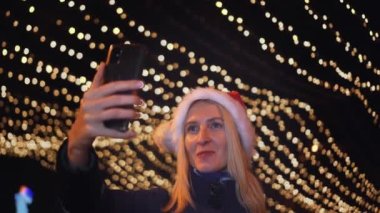 Noel Baba şapkalı güzel sarışın cep telefonuyla konuşuyor ve iyi haberlere duygusal tepki veriyor. Akıllı telefonuna gece şehrin ışıklarının arka planında bakıyor..