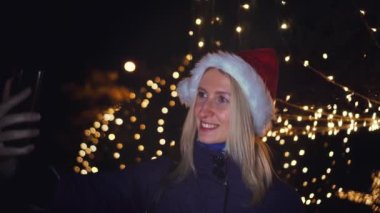 Noel Baba şapkalı güzel sarışın cep telefonuyla konuşuyor ve iyi haberlere duygusal tepki veriyor. Akıllı telefonuna gece şehrin ışıklarının arka planında bakıyor..