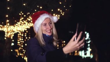 Noel Baba şapkalı güzel sarışın cep telefonuyla konuşuyor ve iyi haberlere duygusal tepki veriyor. Akıllı telefonuna gece şehrin ışıklarının arka planında bakıyor..