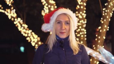 Makyajlı bir kadın portresi, kameraya gülümseyen ve gece veya akşam şehir ışıklarının arka planında Noel Baba şapkası takan. Gülümser, yeni yılı ya da Noel 'i kutlar..