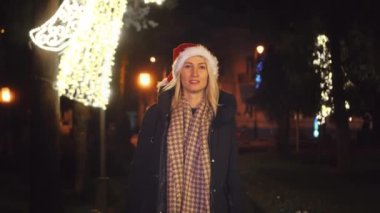 A beautiful woman in a Santa Claus hat is walking on a winter night. The street is decorated with garlands of flowers along the alley, decorated with lights on New Year's Eve or Christmas.