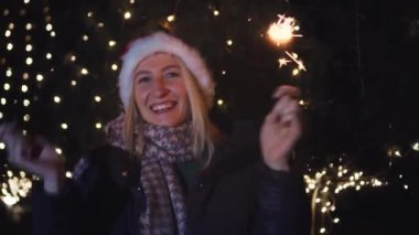 Kameraya gülümseyen ve arka planda gece veya akşam ışıkları karşısında Noel Baba şapkası takan bir kadın. Gülümsüyor, seviniyor, elinde alevler yanıyor, Noel 'i kutluyor..