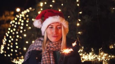 Sarışın bir kadın yılbaşını ya da Noel Baba şapkasıyla kutluyor. Arka planda şehir ışıkları ve çelenkler var. Huysuz, tatminsiz bir kadın üzgün görünüyor, elinde bir maytap yanıyor..