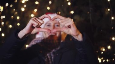 Genç, güzel, beyaz bir kadın Noel 'i kutluyor. Gülümsüyor ve elleriyle kalbinin şeklini gösteriyor. Arka planda, gece kenti veya parkın Noel çelenkleri ve ampulleri yanıyor.