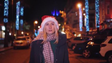 A beautiful woman in a Santa Claus hat is walking on a winter night. The street is decorated with garlands of flowers along the alley, decorated with lights on New Year's Eve or Christmas.
