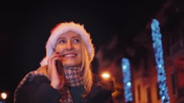 Beyaz bir kadının akıllı telefonuyla sohbet edişinin yakın çekimi. Noel Baba şapkalı güzel bir kadın. Bir kız telefonda konuşuyor ve gülümsüyor. Noel 'de ya da Yeni Yıl' da şehir ışıklarının arka planında..