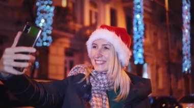 Noel 'de genç ve güzel bir kız gece şehirde dolaşıyor ve selfie çekiyor. Noel ışıkları. Noel Baba şapkası takıyor ve sosyal medya için akıllı telefonuyla fotoğraf çekiyor..