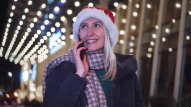 Beyaz bir kadının akıllı telefonuyla sohbet edişinin yakın çekimi. Noel Baba şapkalı güzel bir kadın. Bir kız telefonda konuşuyor ve gülümsüyor. Noel 'de ya da Yeni Yıl' da şehir ışıklarının arka planında..
