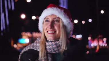 Beyaz bir kadının akıllı telefonuyla sohbet edişinin yakın çekimi. Noel Baba şapkalı güzel bir kadın. Bir kız telefonda konuşuyor ve gülümsüyor. Noel 'de ya da Yeni Yıl' da şehir ışıklarının arka planında..