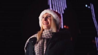 close-up of a white woman chatting on her smartphone. A pretty woman in a Santa hat. a girl is talking on the phone and smiling against the background of city lights on Christmas or New Year's Day.