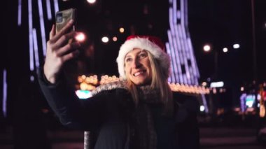 Noel Baba şapkalı güzel sarışın cep telefonuyla konuşuyor ve iyi haberlere duygusal tepki veriyor. Akıllı telefonuna gece şehrin ışıklarının arka planında bakıyor..