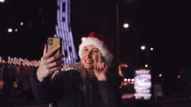 Noel Baba şapkalı güzel sarışın cep telefonuyla konuşuyor ve iyi haberlere duygusal tepki veriyor. Akıllı telefonuna gece şehrin ışıklarının arka planında bakıyor..