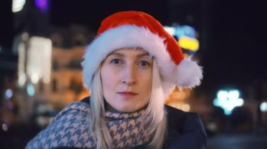 Genç, güzel, beyaz bir sarışın gece şehrinin caddesindeki kameraya bakıyor. Noel Baba şapkası takıyor. Kızım. Yeni yıl. Noel tatili konsepti