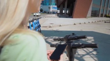 Modern şehrin elektrikli scooter 'ının yanında telefonu tutan kadınların yakın çekimleri, kilidi açmak üzere. Modern şehir ulaşımı. Şehir merkezinde eko-ulaşım kullanıyor. Arkadan görünüm.