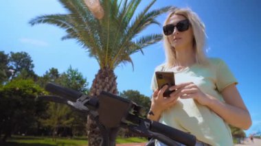 Woman puts smartphone to modern urban electric scooter, intending to unlock it. Modern urban transport. uses eco-transport in city center. palm trees, resort. party or weekend at sea.
