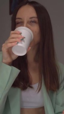 Yeşil fransız ve beyaz bluzlu gülümseyen genç bir kadının portresi, bir fincan kahve ya da çayla birlikte masada oturuyor. Gülümse. İnsanların yaşam tarzı kavramı. Dikey video.