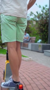Beyaz tişörtlü ve yeşil şortlu genç adam elektrikli scooter ya da scooter kullanıyor. Park ve kaldırım boyunca şehir ve arabaların arka planına karşı dikey video çekiyor. Ağır çekim. arka plan.