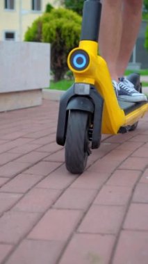 Erkeklerin ayaklarına spor ayakkabılarla yakın çekim, şehrin sokaklarında ve parkta elektrikli scooter ya da scooterla gezinme. Dikey video, yavaş çekim. Sarı ve siyah. Kentte ekolojik ve mobil taşımacılık.