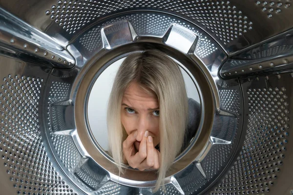 Young blonde woman looks into the drum of a washing machine and holds her nose against an unpleasant smell, photo from the inside