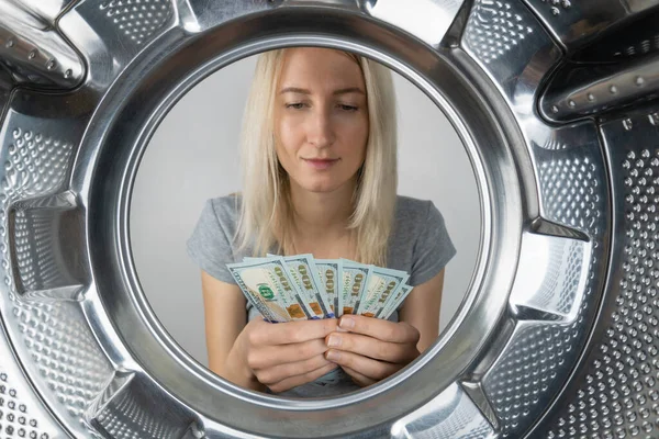 Young woman holds money in her hands near the washing machine, a photo from inside the drum of the washing machine. Concept of payment, money laundering.