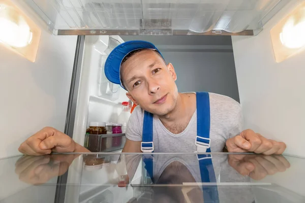 Portrait of a male repairman in uniform inside a refrigerator. Concept of repair of household appliances. Photo from inside the refrigerator