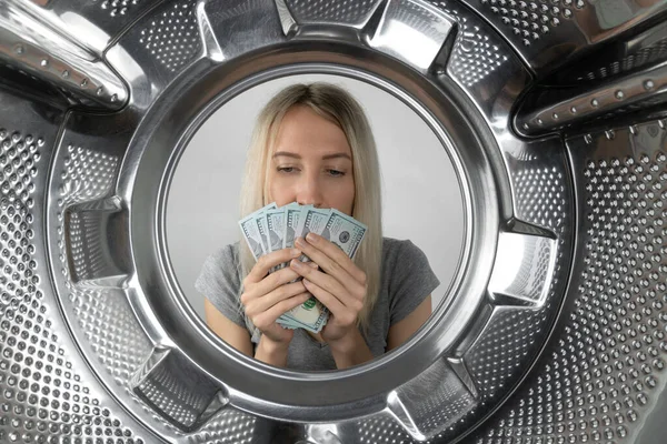 Young woman holds money in her hands and sniffs it near the washing machine, a photo from inside the drum of the washing machine. Concept of payment, money laundering.