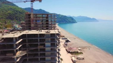 Drone view of construction site, multi-storey buildings under construction against backdrop of sea and mountains on sunny summer day. Construction equipment, crane and workers walk around building.